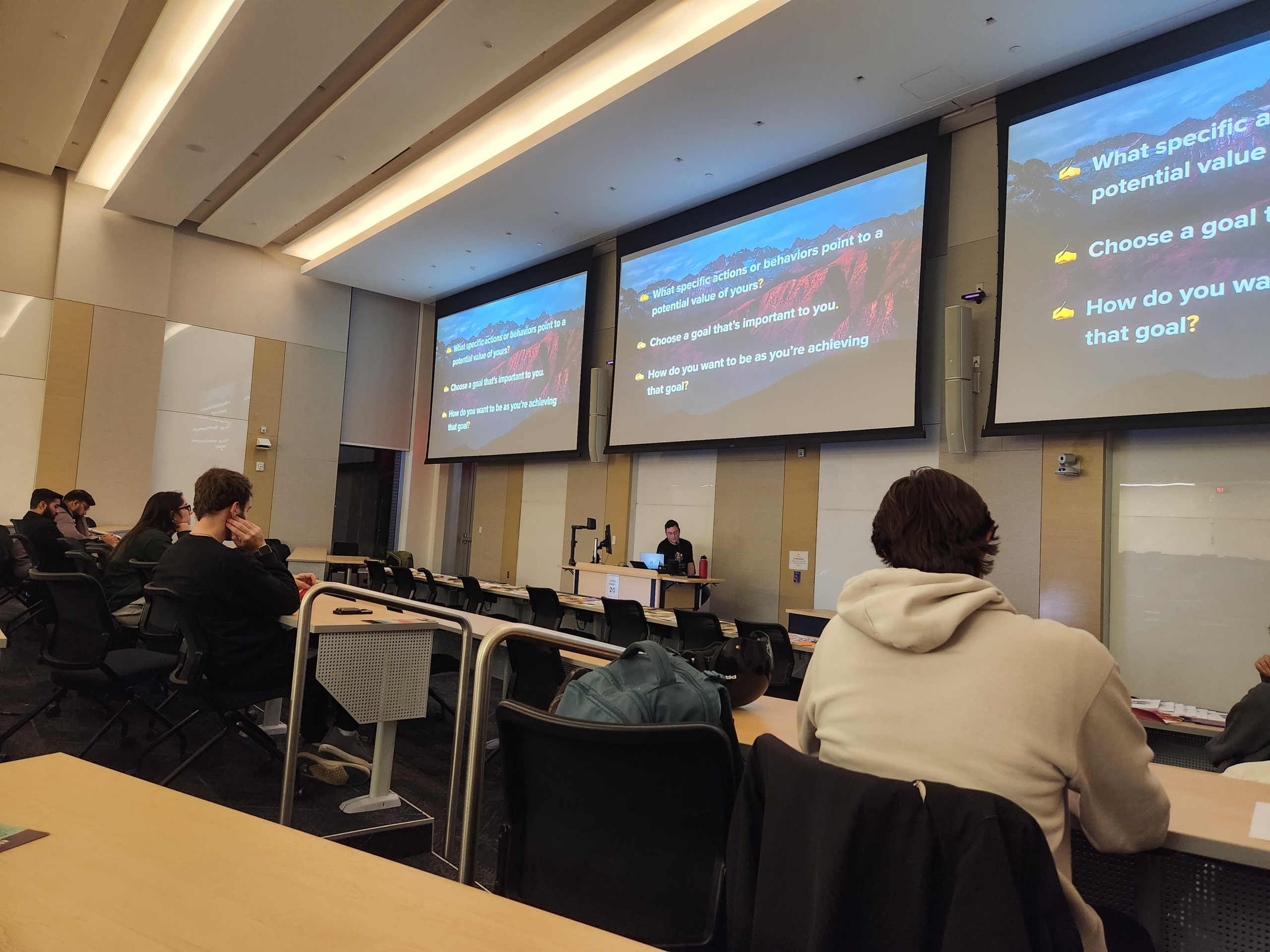 A lecture hall with students facing a presentation screen displaying bullet points about setting goals and determining personal values.