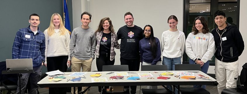 A group of eight people at Fairfield University stands smiling behind a table with various colorful papers and cards, in a room with large windows and overhead lighting.