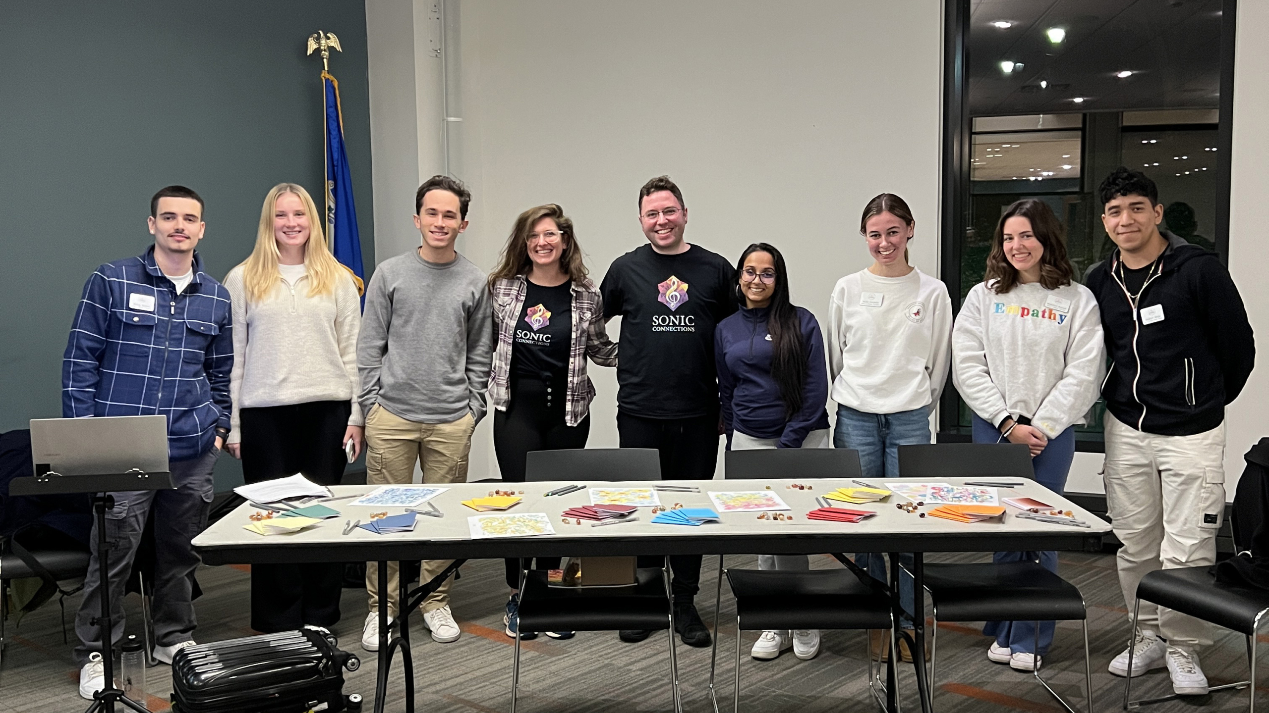 A group of eight people at Fairfield University stands smiling behind a table with various colorful papers and cards, in a room with large windows and overhead lighting.