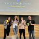 Four people pose with smiles, holding colorful journals in front of a presentation screen that reads "Welcome to... SONIC CONNECTIONS at Wheaton College."