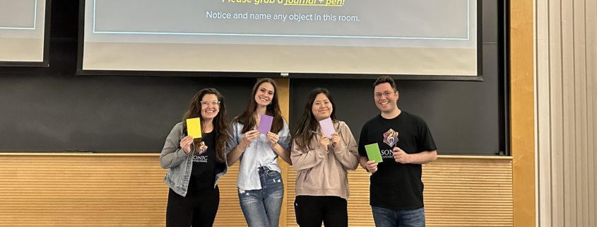 Four people pose with smiles, holding colorful journals in front of a presentation screen that reads "Welcome to... SONIC CONNECTIONS at Wheaton College."