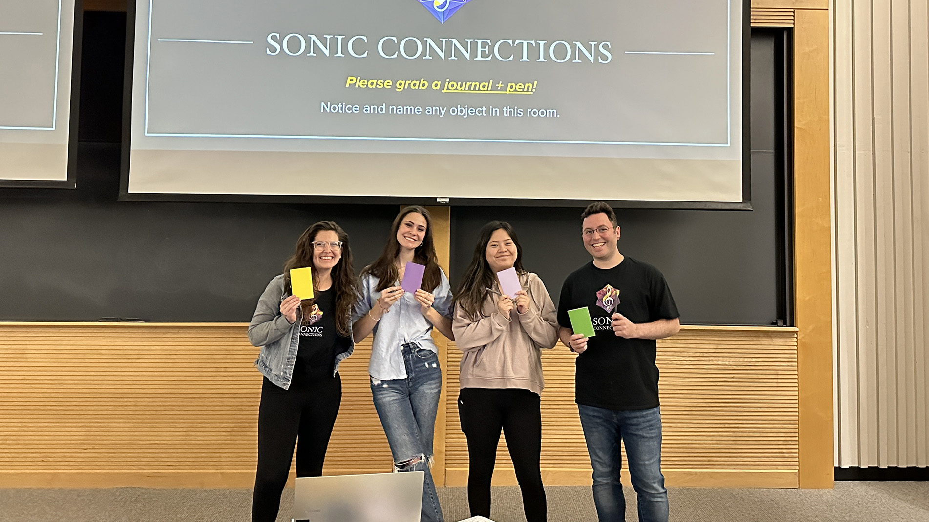 Four people pose with smiles, holding colorful journals in front of a presentation screen that reads "Welcome to... SONIC CONNECTIONS at Wheaton College."