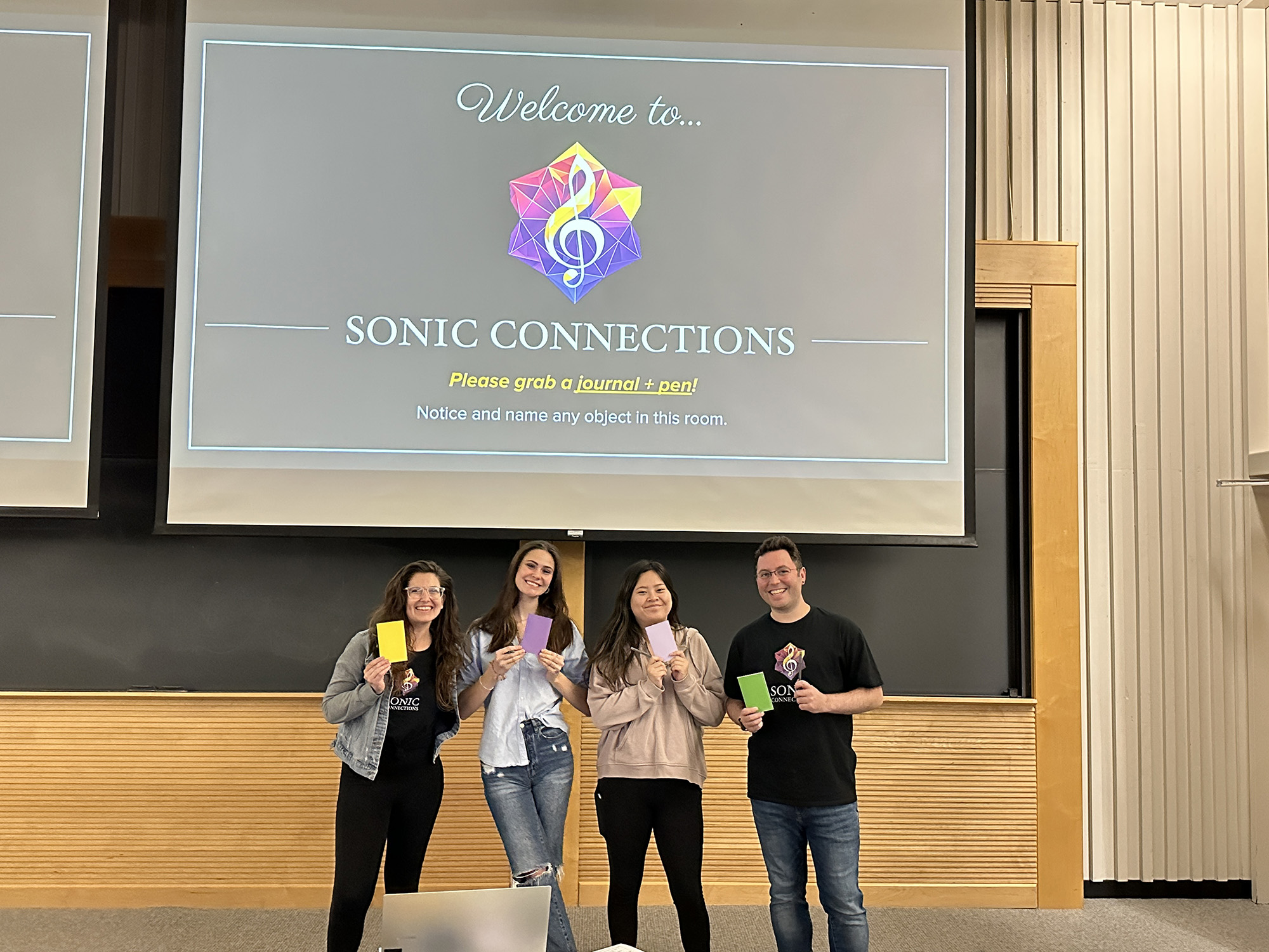 Four people pose with smiles, holding colorful journals in front of a presentation screen that reads "Welcome to... SONIC CONNECTIONS at Wheaton College."
