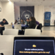 In a Yeshiva University room adorned with "Welcome" banners, a group of people is engaged in an activity, with a laptop screen in the foreground posing the question, "On your wellness journey... What challenges/barriers get in the way?"