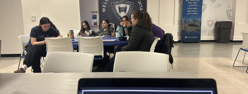 In a Yeshiva University room adorned with "Welcome" banners, a group of people is engaged in an activity, with a laptop screen in the foreground posing the question, "On your wellness journey... What challenges/barriers get in the way?"