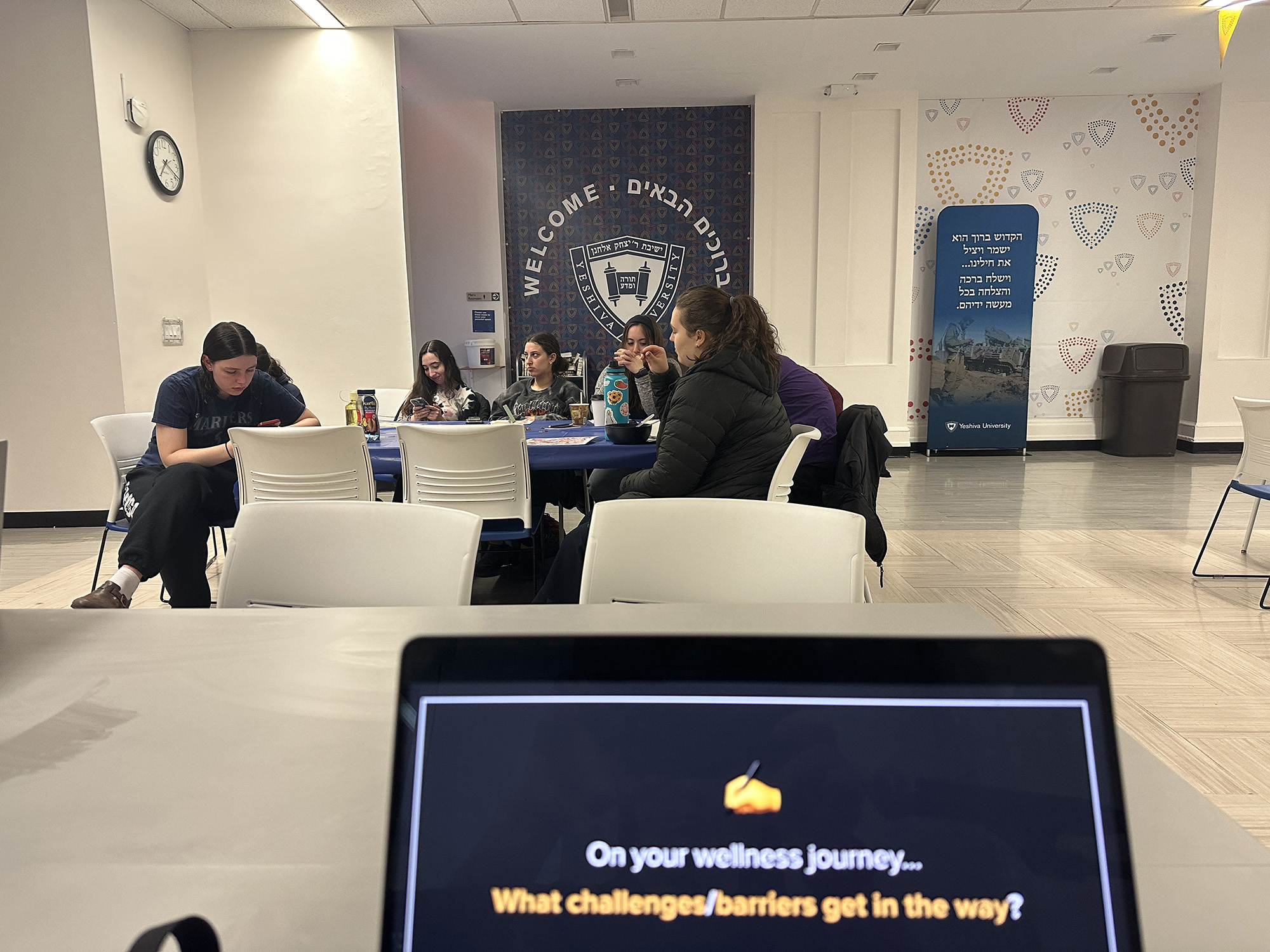 In a Yeshiva University room adorned with "Welcome" banners, a group of people is engaged in an activity, with a laptop screen in the foreground posing the question, "On your wellness journey... What challenges/barriers get in the way?"