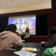 A presenter discusses a colorful, symbolic illustration during a workshop in a hall filled with attending students.