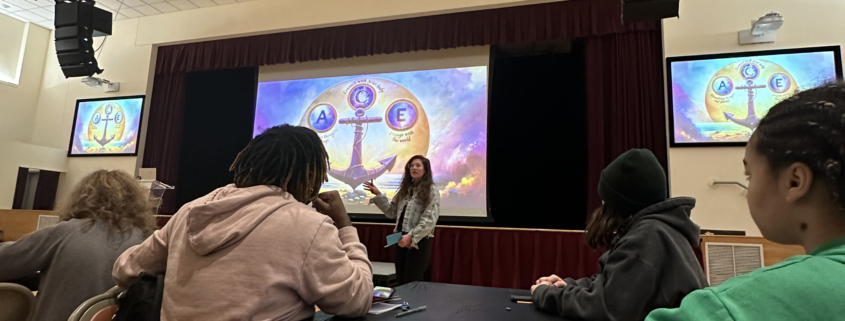 A presenter discusses a colorful, symbolic illustration during a workshop in a hall filled with attending students.